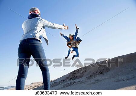 Stock Image - Young woman watching friend jumping above sand dune. Fotosearch