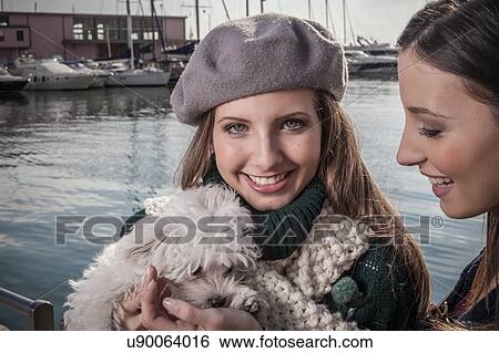 Stock Photograph - Young woman wearing beret in boatyard holding dog looking at camera smiling. Fotosearch