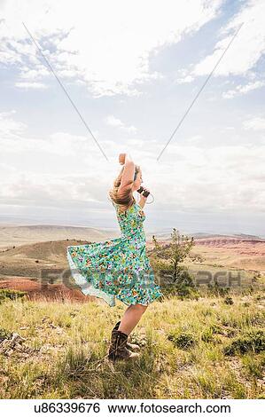 Stock Photograph - Young woman wearing dress and cowboy boots dancing in landscape, Bridger, Montana, USA. Fotosearch