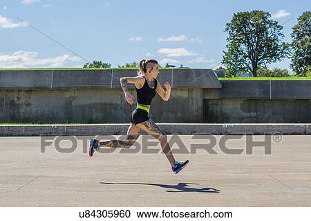 Stock Image - "Young woman with tattoos, running on track, outdoors". Fotosearch