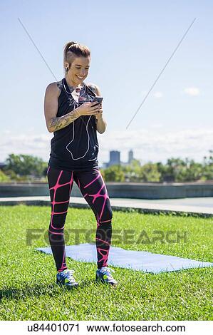 Stock Image - "Young woman with tattoos, using smartphone, wearing earphones, outdoors". Fotosearch