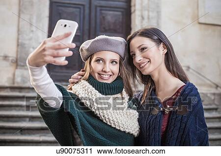 Stock Image - Young women using smartphone to take selfie smiling. Fotosearch