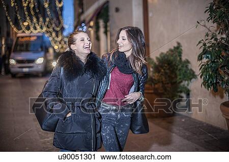 Stock Image - Young women walking down street in the evening arm in arm laughing. Fotosearch