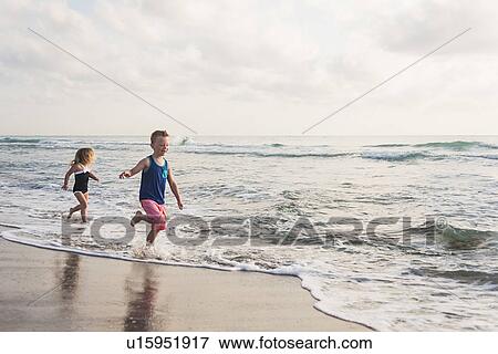 Stock Photo - Boy (6-7) and girl (4-5) running on beach by water. Fotosearch