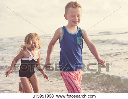 Stock Photo - Boy (6-7) and girl (4-5) running on beach by water. Fotosearch