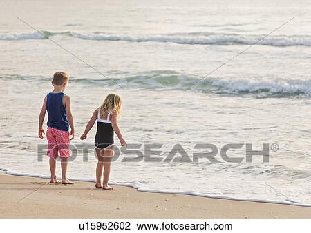 Stock Image - Boy (6-7) and girl (4-5) standing on beach by water. Fotosearch
