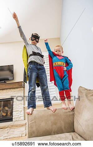 Stock Image - Boys (2-3, 6-7) wearing superhero costumes standing on sofa. Fotosearch