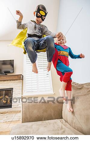 Stock Image - Boys (2-3, 6-7) wearing superhero costumes jumping on sofa. Fotosearch