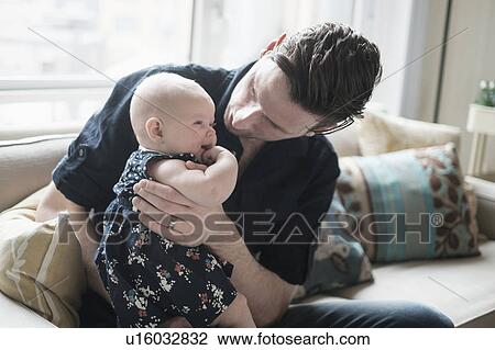 Stock Image - Father sitting on sofa and playing with baby daughter (2-5 months). Fotosearch