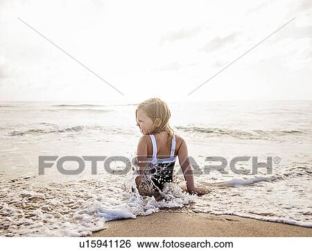 Stock Photograph - Girl (4-5) sitting in water on beach. Fotosearch