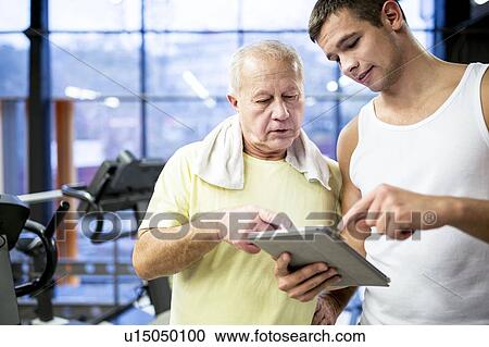 Stock Image - Gym trainer showing tablet to senior man. Fotosearch
