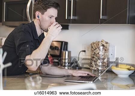 Stock Image - Man drinking while working on laptop computer in kitchen. Fotosearch