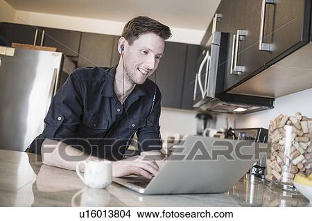 Picture - Man smiling while working on laptop computer in kitchen. Fotosearch