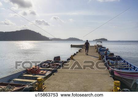 Stock Image - Man walking along pier with boats moored along. Fotosearch