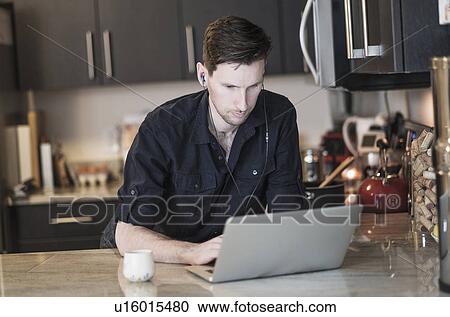 Stock Image - Man working on laptop computer in kitchen. Fotosearch