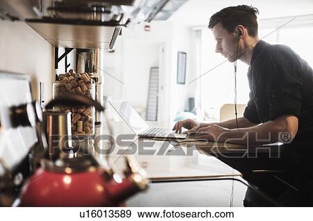 Stock Photo - Man working on laptop computer in kitchen. Fotosearch