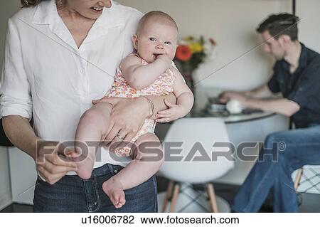 Stock Image - Mother holding baby (2-5 months) and father working in background. Fotosearch