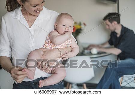 Mother holding baby (2-5 months) and father working in background View Large Photo Image Stock Photo - Mother holding baby (2-5 months) and father working in background. Fotosearch