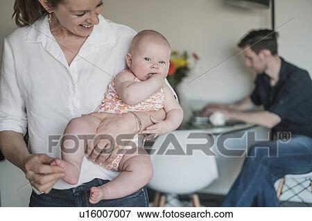 Stock Photo - Mother holding baby (2-5 months) and father working in background. Fotosearch