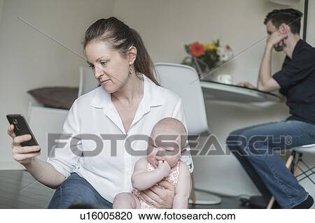 Mother holding baby (2-5 months) while using smart phone and father working in background View Large Photo Image Stock Image - Mother holding baby (2-5 months) while using smart phone and father working in background. Fotosearch