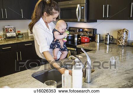 Stock Image - Mother holding baby daughter (2-5 months) and filling glass with water. Fotosearch