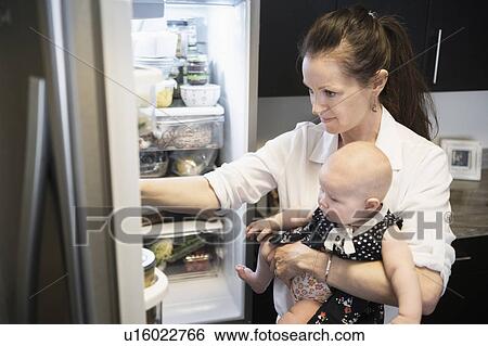 Stock Photograph - Mother looking for food in refrigerator while holding baby daughter (2-5 months). Fotosearch
