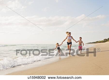 Stock Image - Mother running with boy (6-7) and girl (4-5) on beach by water. Fotosearch