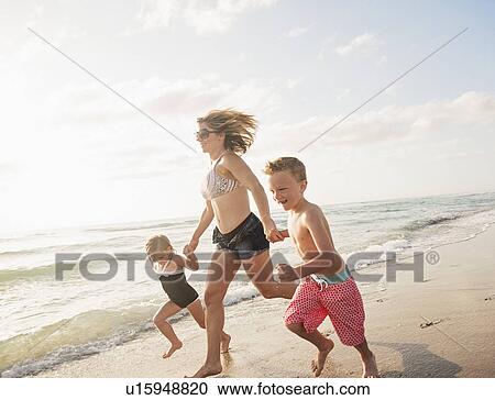 Stock Image - Mother running with boy (6-7) and girl (4-5) on beach by water. Fotosearch
