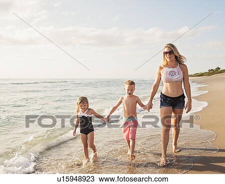 Stock Image - Mother walking with boy (6-7) and girl (4-5) on beach by water. Fotosearch