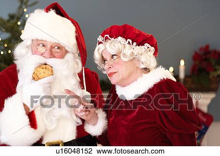 Stock Image - Mrs. Claus pointing on Santa eating gingerbread cookie. Fotosearch