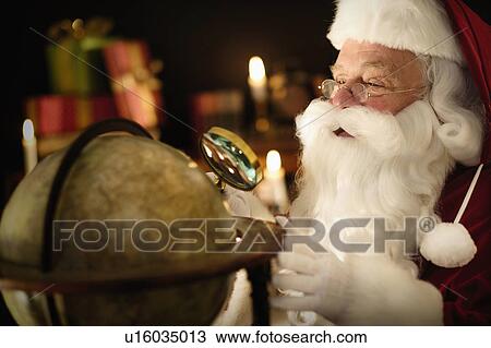 Stock Image - Santa Claus looking through magnifying glass at globe. Fotosearch