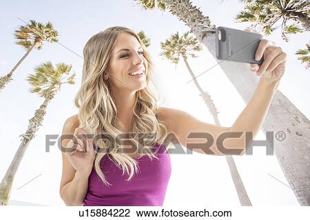 Stock Image - Smiling blonde woman taking selfie against palm trees. Fotosearch