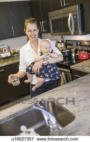 Stock Photo - Smiling mother holding baby daughter (2-5 months) and glass of water. Fotosearch