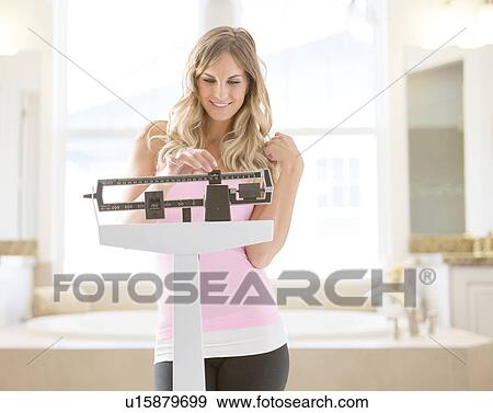 Stock Photo - Young woman standing on weight scale in bathroom. Fotosearch