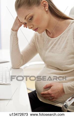 Stock Image - Pregnant woman at desk with head in hands. Fotosearch
