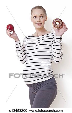 Stock Image - Pregnant woman holding apple and doughnut. Fotosearch