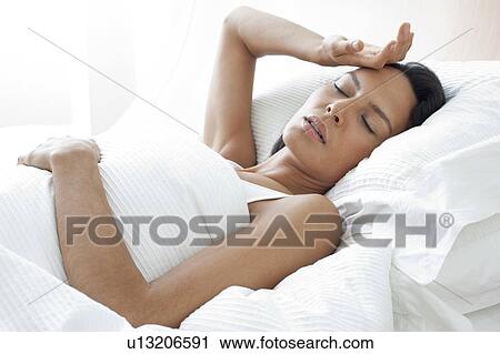 Stock Image - Young woman in bed with hand on forehead. Fotosearch