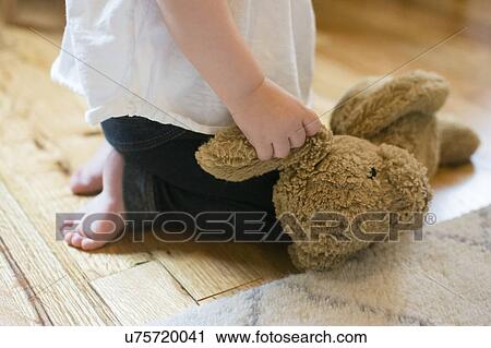 Stock Image - Baby girl (18-23 months) kneeling on floor and holding teddy bear. Fotosearch
