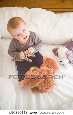 Stock Photo - Baby girl sitting on bed holding soft toys looking up at camera. Fotosearch