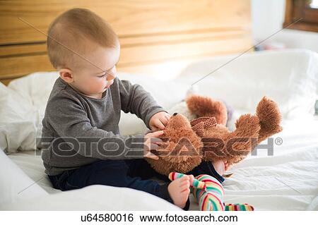 Stock Photograph - Baby girl sitting on bed playing with teddy bear. Fotosearch