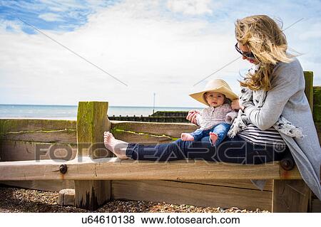Stock Photo - Baby girl wearing sun hat sitting on groyne with mother. Fotosearch