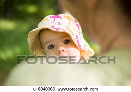 Stock Photograph - Baby girl wearing sunhat looking over mothers shoulder at camera. Fotosearch