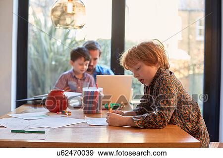 Stock Photo - Boy colouring at dining table whilst father using laptop with brother. Fotosearch