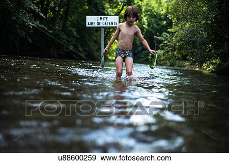 Stock Photo - Boy knee deep in water holding shoe, France. Fotosearch