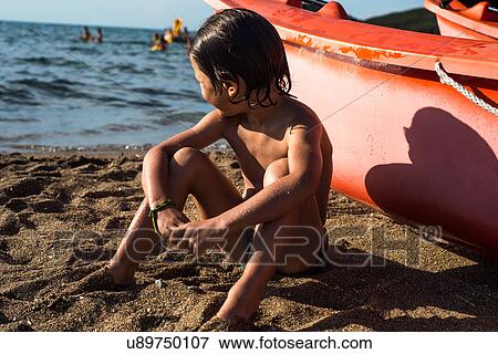Stock Photo - Boy sitting on beach by boat looking away. Fotosearch