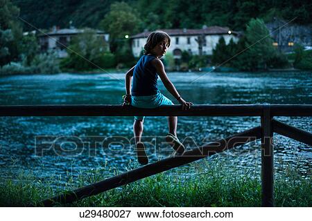 Stock Photo - Boy sitting on fence by river looking over shoulder at camera. Fotosearch