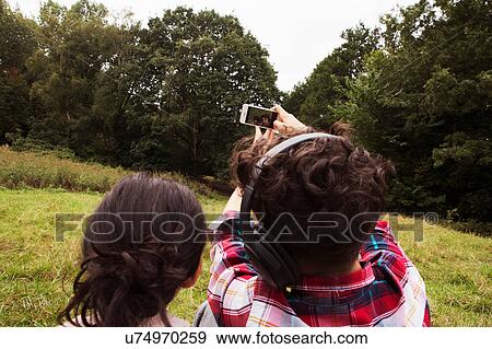 Stock Photo - Brother and sister, sitting outdoors, taking selfie, using smartphone, rear view. Fotosearch