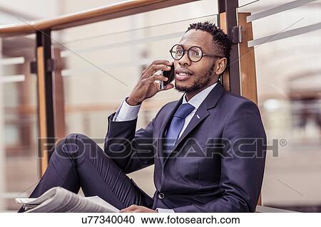 Stock Image - Businessman sitting on floor chatting on smartphone on office balcony. Fotosearch