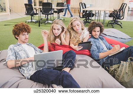 Stock Image - Co-workers working on laptop on bean bags in open plan office. Fotosearch