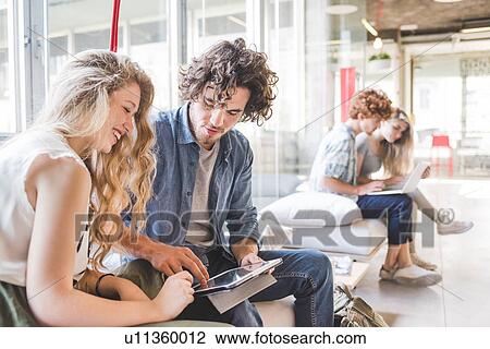 Stock Image - Co-workers working on laptop on sofa by window. Fotosearch